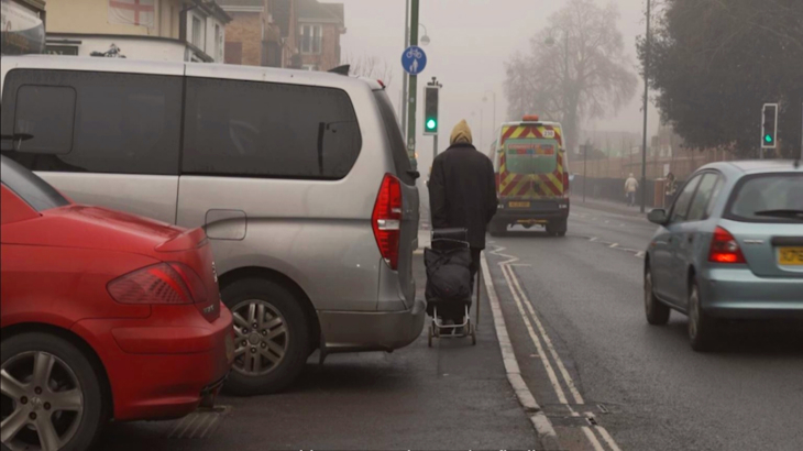 A van shown parked over a pavement on a street on a misty day ahead a person with a trolley is walking along the pavement