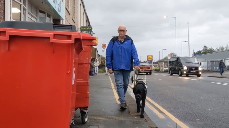 Mark and his guide dog Bobby walking towards the camera on a narrow pavement partially obstructed by large red bins, with traffic going past