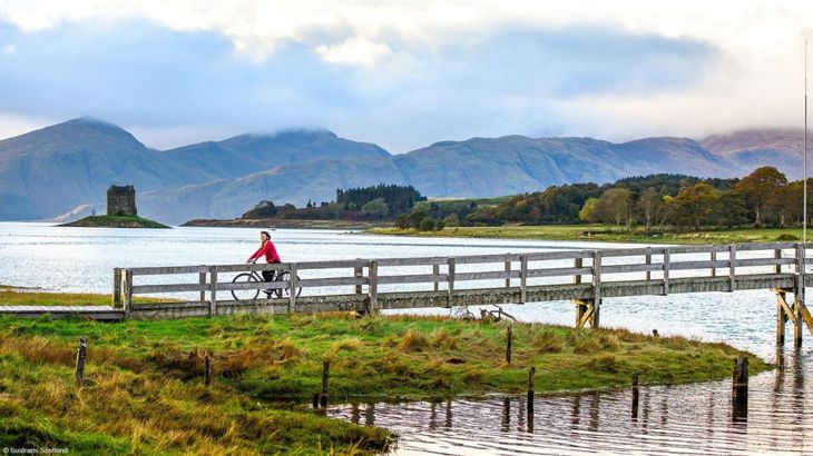 A woman rides a bike over a bridge on the Caledonia Way, with amazing landscape and a castle in the background.