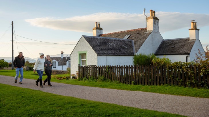 3 adult pedestrians at Crinan, NCN 78, The Caledonia Way. They are wearing warm clothes and walking along the path past a white house behind them.