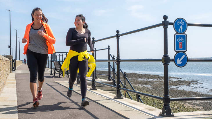 Two women jogging along a coastal path in Broughty Ferry, Dundee wearing luminous clothing. A beach and the North Sea in the background.