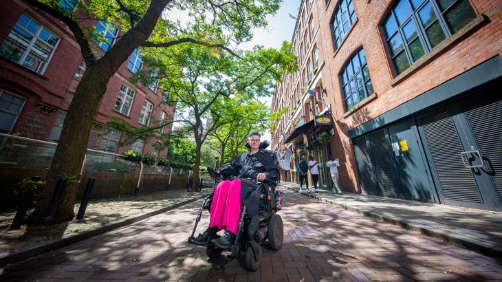 A person with short hair and bright pink trousers on in an electric wheelchair in the middle of Manchester's city centre in the summer time