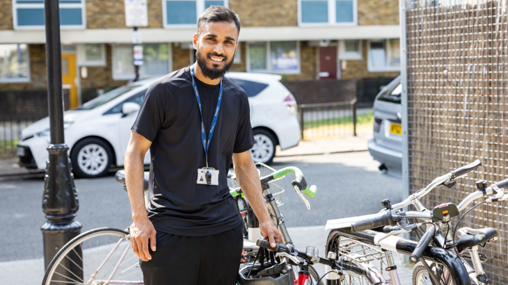 A male school teacher stood smiling in the sunshine in Tower Hamlets in London holding a bike and wearing a lanyard and a whistle around his neck
