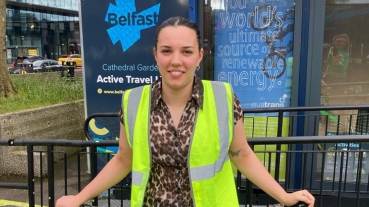 A woman wearing a hi-vis vest stands in front of Cathedral Gardens Active Travel Hub in Belfast city centre.