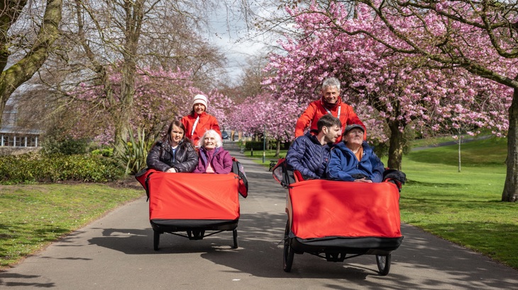 Participants are shown using E-trishaws during a Cycling without Age ride. There are two E-trishaws, each with a rider and two people sat in front.