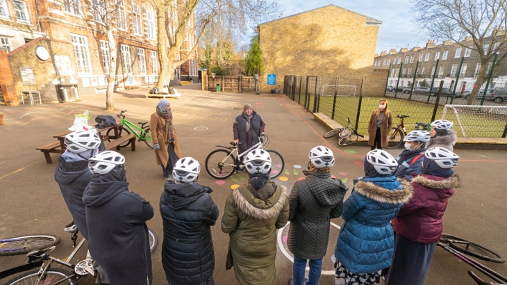 A group of mothers standing beside bicycles take part in a cycling workshop led by instructor. They are in the grounds of a school.
