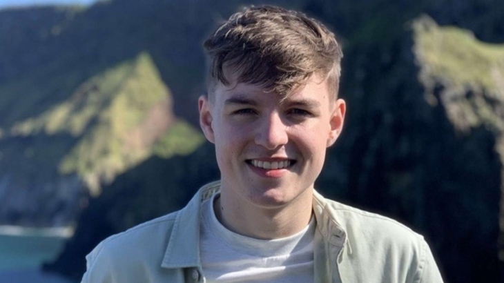 A young man smiles for the camera from a scenic spot beside sea cliffs on a bright sunny day.