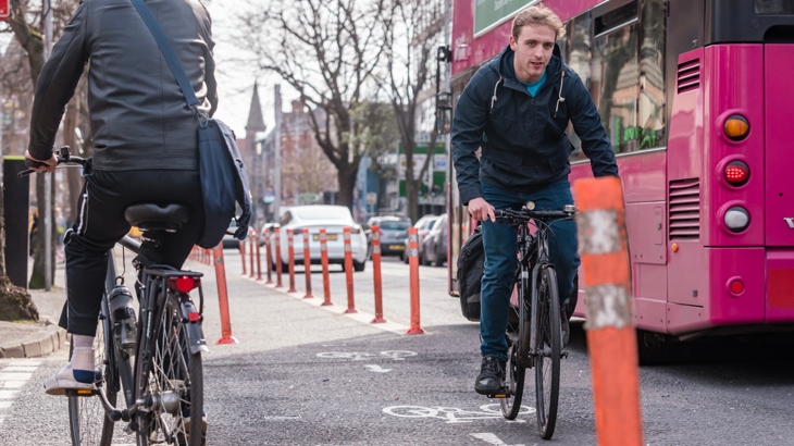 A man on a bike passes another cyclist along a cycle lane while a double-decker bus passes close by.