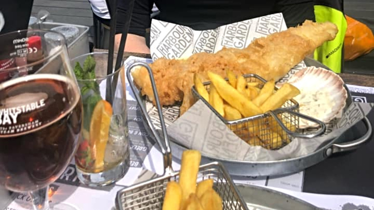 A plate of fish and chips on an outdoor table next to a glass of beer on a sunny day
