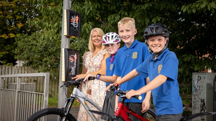 A woman and three school pupils stand at a road crossing, two of the children have bikes and are wearing helmets.