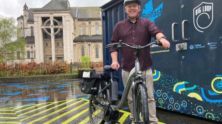 A man wearing a helmet stands with an electric bicycle beside a cycle storage container with a cathedral in the background.