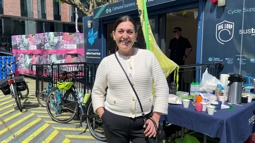 A woman with dark hair wearing an 'I heart cycling' badge on her cream cardigan smiles for the camera outside Cathedral Gardens hub.