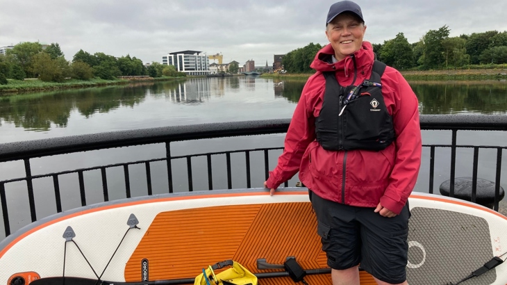 A woman wearing a red coat, cap and black life-jacket stands with an orange paddleboard on a bridge over the River Lagan in Belfast.