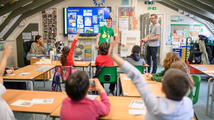 Two Walk Wheel Cycle Trust staff members stand in front of a classroom of children, some of whom have their hand up