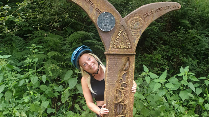 A woman with blonde hair smiling while peering around a cast-iron sculpture called a Millennium Mileposts on the Network