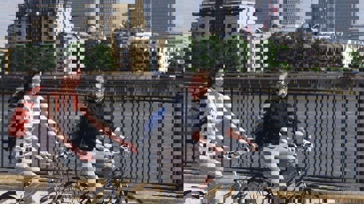 Two Women Cycling Along The Thames With Canary Wharf In The Background