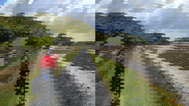 Cyclist in red jacket on gravel path by estuary with green hills and trees to the side
