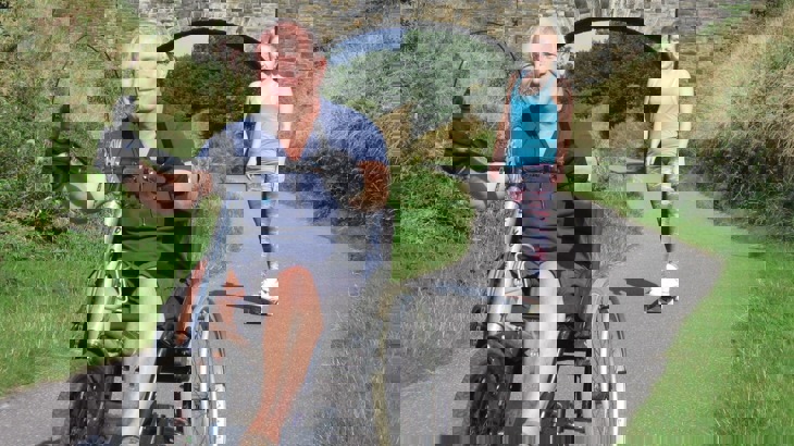 A man on a hand tricycle and girl on a skateboard on tarmac greenway