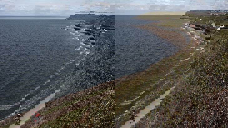 Two cyclists on coastal cycle path with sea wall and beach and buildings in the distance