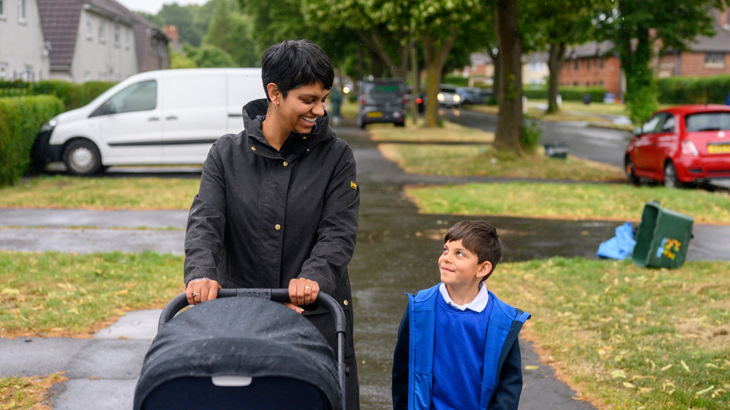 A woman pushing a pram in a residential area on a rainy day smiling and looking down at a small child wearing a school uniform who is looking up and smiling back