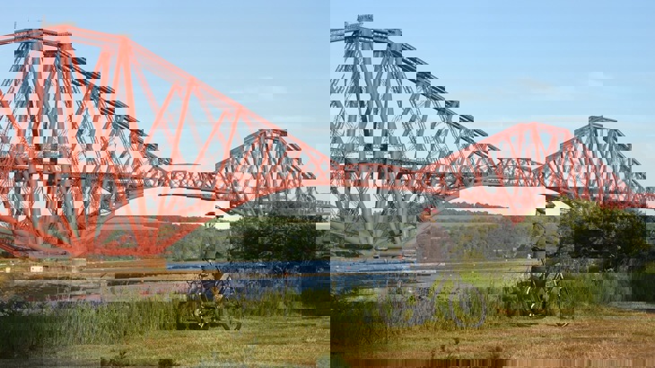 Cyclist in helmet and grey hoodie standing on grassy riverbank, looking at large, red metal bridge