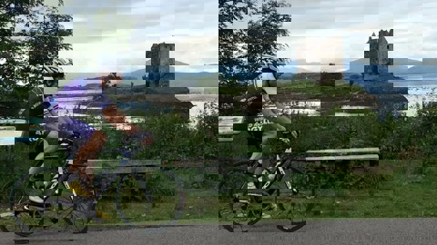 Man cycling on road bike on asphalt path in white helmet and blue and white lycra. To the side is loch with island and castle on it