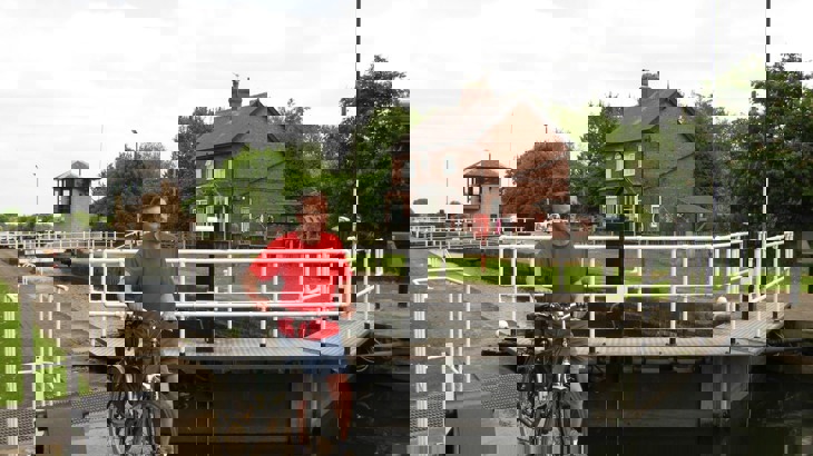 Man in t shirt and shorts pushing bike at side of canal locks with red brick house in the background