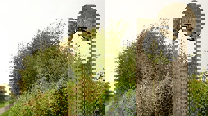 Flat wooden sculpture with circular hole in front of bushes, next to cycle path