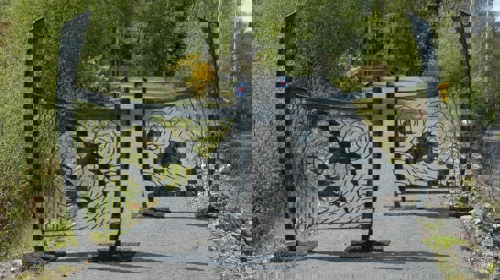 A pair of decorative metal panels, staggered on a traffic-free path to control access. The curved panels have silhouette images on them, one has two birds in flight, the other a puffing steam train. The path beyond continues on uphill where the path enters a wooded area. An interpretation board is in the distance.