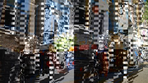 A man stands astride a cargo bike loaded with shopping. He is outdoors on a London street. He is smiling, the weather is fine and the mood is pleasant.