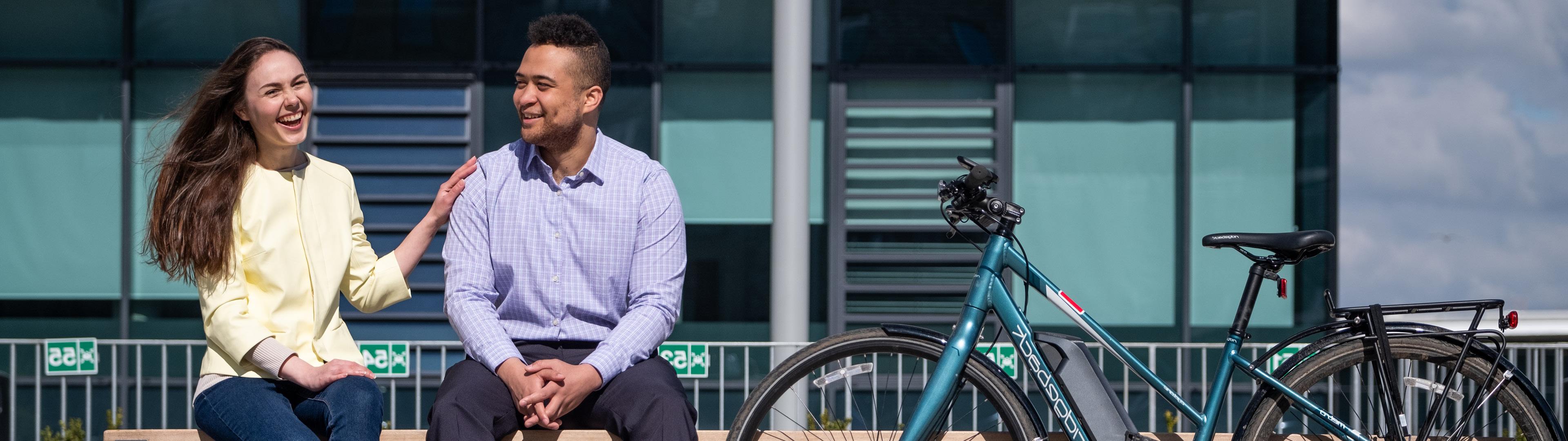 Man and woman sitting on a bench chatting and laughing, with a bicycle propped up next to them.