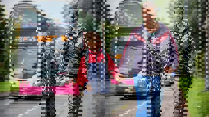 Child looks up at parent as they walk to school along the pavement with buses driving past