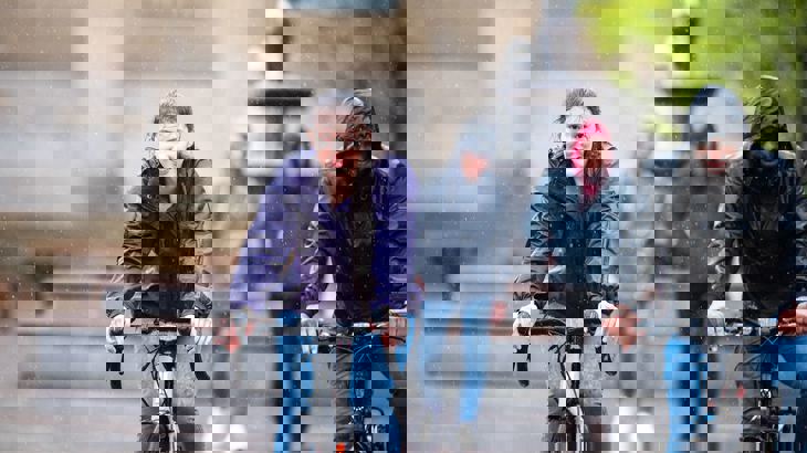 Lady wearing a purple waterproof smiling as she cycles in the pouring down rain next to her male friend in a black waterproof jacket with his hood up.