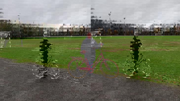 Jane, white female, stands with her maroon Dutch-style cycle on a gravel path on the edge of a sports field. Jane has cropped white hair and is wearing a black coat and blue jeans. There are several football posts in the field behind her, and trees in the distance.
