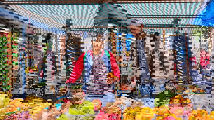 A man and woman holding their bikes, chatting as they buy fruit at a food market in London.