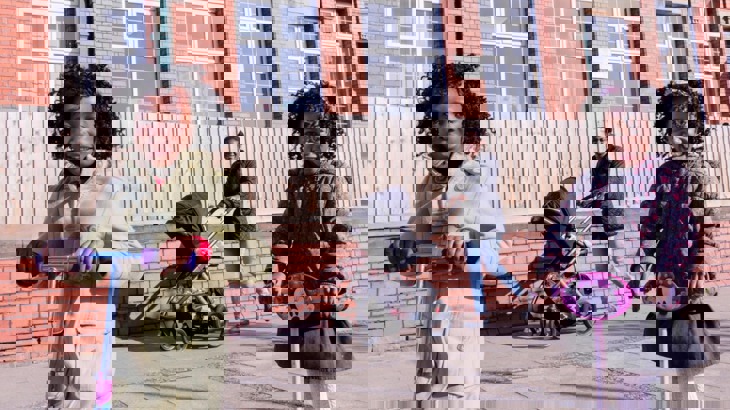 Two young girls on their scooters smile at the camera. Their mum is walking behind with a pushchair