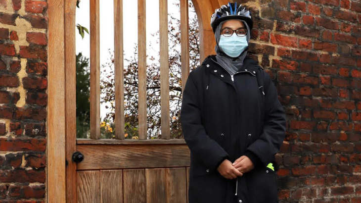 Shaima standing in front of a wooden doorway in a local park, wearing a protective mask and a cycle helmet.