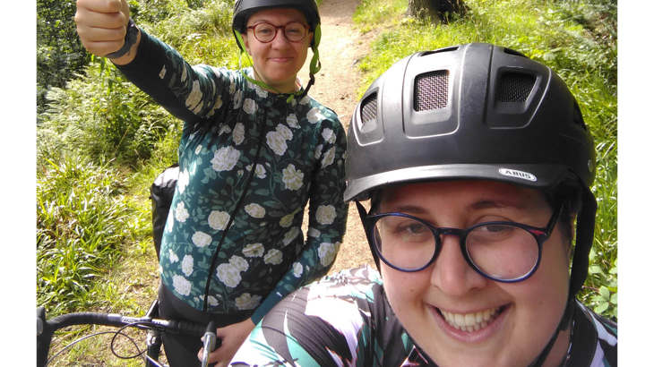 Seflie with Abi and Lilith, smiling with their thumbs up, wearing cycling helmets and standing with their bikes in a green park.