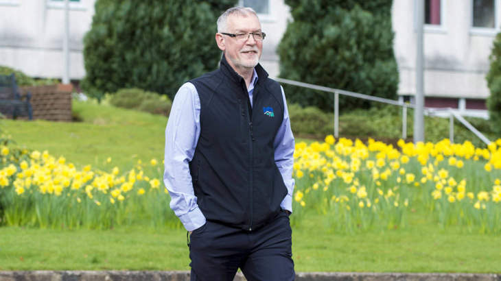 Chief Officer At Paths For All, Ian Findlay, smiling as he walks through a green park surrounded by daffodils