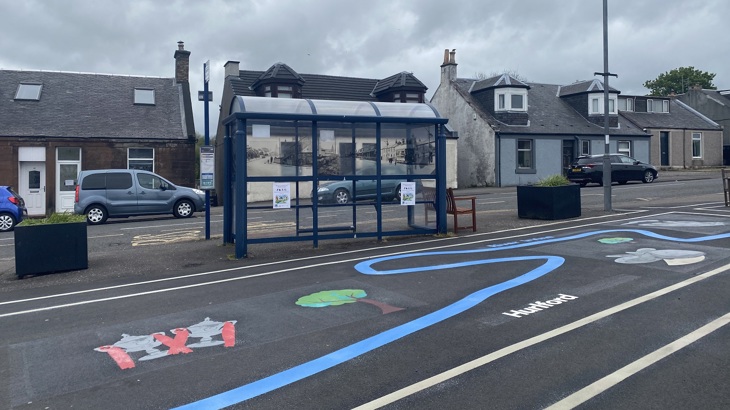 A bus stop in the background with artwork on the ground, planters and benches, part of a pop-up parklet.