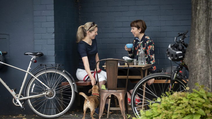 Two women chat at an outdoor cafe with bikes parked nearby. One has a dog on lead. 