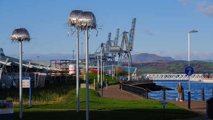 Tragic O'Hara's sculpture Mechanical Animals at Greenock Waterfront. Three large, perspex and metal jellyfish on top of recycled telephone poles beside the River Clyde, with shipyards in the background.