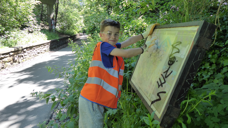 A young person, wearing an orange hi-vis vest, uses a graffiti removal wipe to clean an interpretation board along a traffic-free shared used path in Edinburgh.