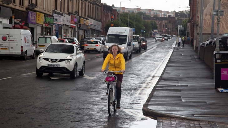 Woman on a bike cycles alongside cars on a busy, wet road with puddles in Glasgow.