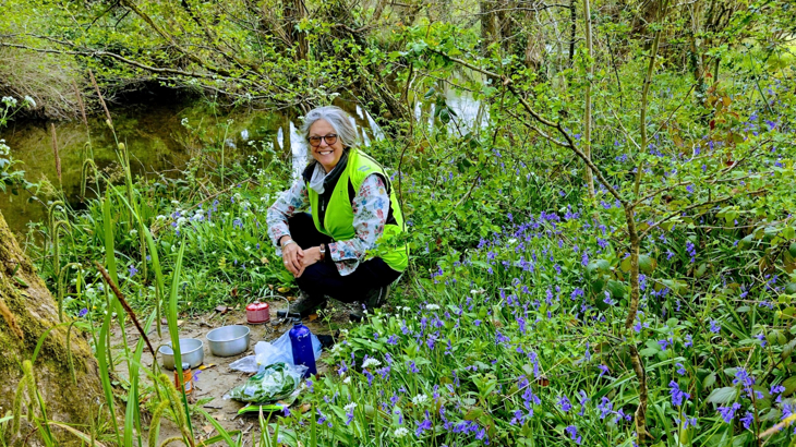 A woman with grey hair kneeling over cooking pots amongst bluebells while smiling at the camera in rural England