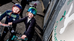 A boy and a girl wearing helmets are on bikes outside their school gates.
