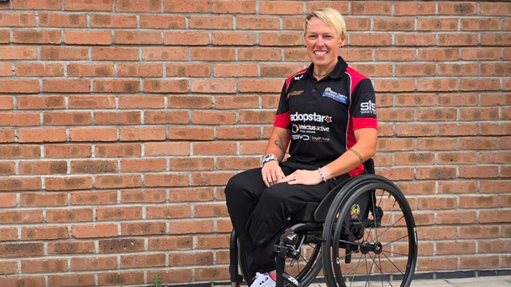 A woman with short blonde hair sat smiling in her wheelchair sat in front of a brick wall outside