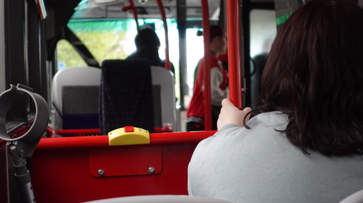 A bus passenger sits near the wheelchair space, holding onto a red rail beside a stop button, with other passengers visible in the background.