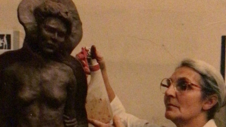 A woman wearing glasses and a white coat with her hair tied back works on a sculpture.