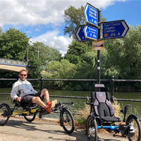 A man sat smiling on an adapted trike next to a river on a sunny day